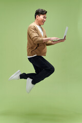 Full length portrait of a happy young man jumping with laptop computer isolated over green background