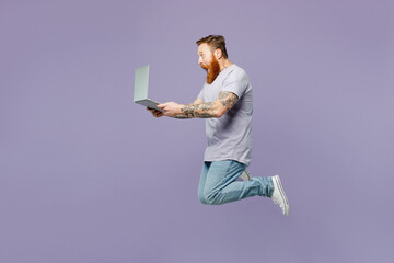 Full body young redhead bearded IT man wear violet t-shirt casual clothes jump high hold use work on laptop pc computer isolated on plain pastel purple background studio portrait. Lifestyle concept.
