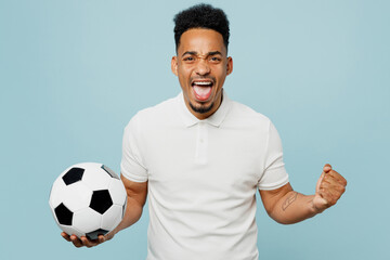 Wall Mural - Young man fan wear basic t-shirt cheer up support football sport team hold in hand soccer ball watch tv live stream do winner gesture clenching fists isolated on plain pastel blue background studio.