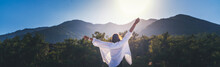 A Woman In A White Shirt With Her Hands Raised Up Enjoy The Morning Sun Against The Backdrop Of Mountains At Sunrise