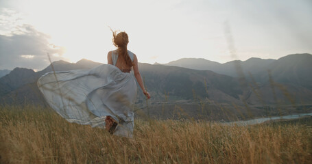 Wall Mural - Young beautiful girl with red hair wearing white dress running on top of a mountain facing wind blowing her hair and dress and smiling - freedom, adventure, harmony 