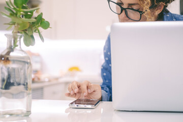 Close up of a woman at home working busy with phone and laptop with white kitchen in background. Modern female people lifestyle and connection. Alternative office and workplace people lifestyle