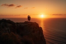 Two People Standing On The Edge Of A Cliff Looking Out At The Sun Setting In The Sky Over The Ocean