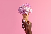 A Female Dark - Skinned Hand With Pink Nails Holding An Ice Cream Waffle Cone With Flowers And Cherry Blossom On A Seamless Pink Background