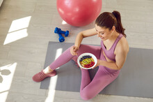 Happy Beautiful Young Woman Having Good Healthy Snack After Workout, Sitting On Yoga Mat On Floor With Fit Ball And Dumbbells, Eating Muesli, Kiwi, Blueberries And Raspberries. Sport Food Diet Concept