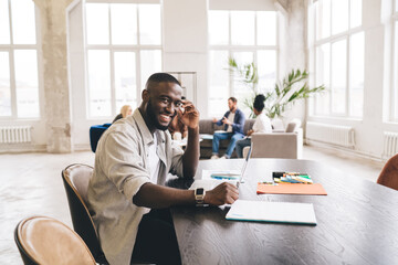 Wall Mural - Cheerful black man sitting at table with laptop and papers