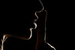 Silhouette of unknown woman , face in the shadow holding finger on her lips on a black background