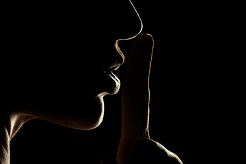 Wall Mural - Silhouette of unknown woman , face in the shadow holding finger on her lips on a black background