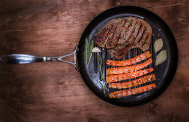 Wall Mural - fried piece of beef on a round frying pan with fried carrots in sesame on a brown wooden background, top view