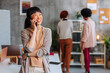 © bernardbodo - Asian business woman talking on phone and laughing while two female coworkers talk in the background.