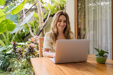 Young woman freelancer working on laptop computer sitting at table on wooden terrace of bungalow  surrounded with greenery