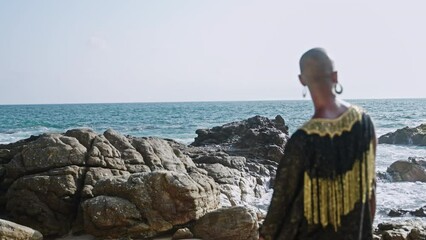 Wall Mural - Queer multiracial model poses at scenic seaside location back view. Non-binary black person in outrageous gown stands with their back to camera at rocky ocean beach. Male gay in posh dress by the sea