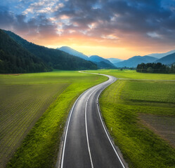 Poster - Aerial view of road in green meadows at sunset in summer. Top drone view of rural road, alpine mountains. Colorful landscape with curved highway, hills, fields, green grass, orange sky. Slovenia