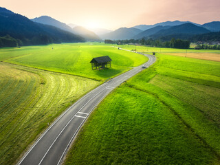 Wall Mural - Aerial view of road in green meadows at sunset in summer. Top drone view of rural road, alpine mountains. Colorful landscape with curved highway, hills, fields, green grass, orange sky. Slovenia