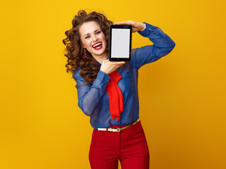 Wall Mural - happy modern woman with long wavy brunette hair against yellow background showing tablet PC blank screen