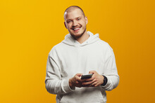 Cheerful Unshaven Man Using Mobile Phone, Wearing Hoodie And Smiling At Camera, Isolated Over Yellow Background