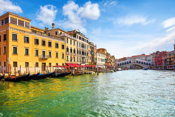 Wall Mural - Venice, Italy. Panorama view at Rialto Bridge on Grand Canal in Venezia. Gondolas floating by piers docksamong antique buildings and traditional italian Venetian architecture. Sunny day with blue sky 