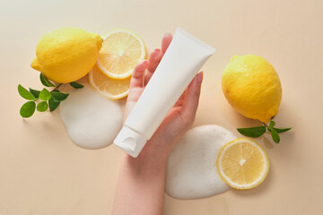 Canvas Print - A white cosmetic tube on female hand with fresh lemon and green leaf on beige background. Blank label for branding mockup. Skincare product concept with natural vitamin C.