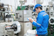 © Nutchapong Wuttisak - back photo American male engineer wearing uniform, hard hat and checking bill of materials with pen It is close to the operating machine control panel. in a large plastic and steel industry