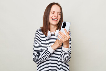 Wall Mural - Smiling laughing woman wearing striped shirt standing with headphones over neck isolated over gray background using mobile phone chatting with friends.