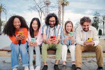 Group of best friends enjoying and smiling using their mobile phone app sitting on a bench sharing messages. Happy young people chatting on social media online with the cellphone having fun together