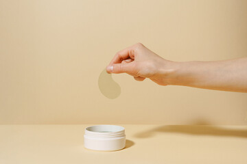 female hand holding sample of green algae extract eye patch over white jar of product on beige isola