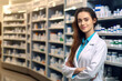 Portrait of young woman pharmacist wearing lab coat standing in pharmacy drugstore and looking at camera.