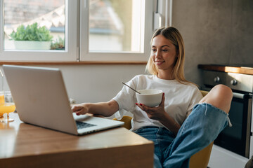 Blond young woman using laptop in the kitchen while having breakfast.