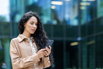 Serious business woman with phone in hands, latin american woman thinking typing message and reading news, boss using app on smartphone while walking outside office building.
