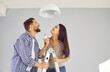© Studio Romantic - Happy family couple changing an energy saving lightbulb at home. Joyful young man and woman standing on a stepladder, holding a LED light bulb, smiling and looking up at the white lamp on the ceiling