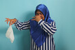 © Jamaludinyusup - A displeased middle-aged Asian woman, wearing a blue hijab, shows disgust as she abominates the smell of used toilet paper and used tissues, while standing against a blue background.'
