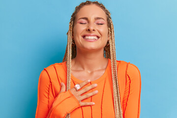 Wall Mural - Close-up of happy young woman with long fair dreads laughing with one hand on her chest, wearing orange top, happy moment concept, copy space