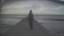 Young Elegant Woman In A Long Black Dress Walking On Pier. View Through Transparent Tulle