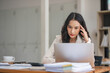 © chartchai - A thoughtful Asian businesswoman focuses on her tasks on her laptop, looking at her laptop with a serious face, and working at her desk in the office.