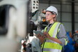 © FotoArtist - Female Automotive Engineer Wearing Hard Hat, Standing, Using Laptop. Monitoring, Control, Equipment Production. Automated Robot Arm Assembly Line Manufacturing Electric Vehicle