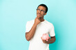 © luismolinero - African American handsome man holding a piggybank over isolated blue background having doubts