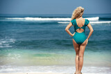 Fototapeta  - A girl in a green swimsuit stands on the beach and looks at the ocean. View of a woman's back against the background of water and sand. Beach holidays in Bali. A blonde with tanned skin enjoys.