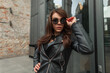 © alones - Fashionable beautiful cool woman model in stylish urban rock black clothes with leather jacket and dress wearing fashion sunglasses in the city near a vintage building