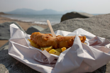 Fish and chips, battered cod, in a tray wrapped in paper with a wooden fork on a beach. Traditional British food concept