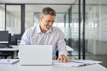 Happy mid aged professional business man company executive ceo manager lawyer sitting at desk in office using laptop working on plan overview checking bills corporate financial account documents.