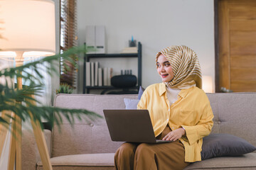 Wall Mural - A stunning Muslim millennial businesswoman wearing a yellow hijab working from her home office while sitting on a couch in her living room.