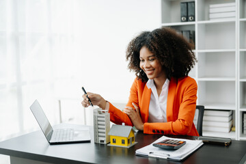 Young real estate agent worker working with laptop and tablet at table in office and small house beside it..