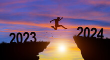 Welcome Merry Christmas And Happy New Year In 2024. Man Jumping Across The Gap From 2023 To 2024 Cliff With Sunset And Twilight Sky Background.