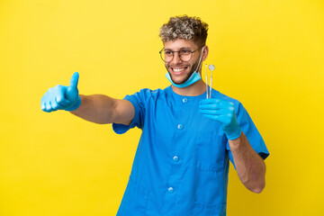 Wall Mural - Dentist caucasian man holding tools isolated on yellow background giving a thumbs up gesture