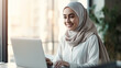 © annebel146 - Portrait of successful Muslim businesswoman inside office with laptop, woman in hijab smiling and looking at camera, muslim office worker wearing glasses.