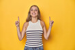 © Asier - Young blonde Caucasian woman in a white tank top on a yellow studio background, indicates with both fore fingers up showing a blank space.