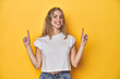 © Asier - Young blonde Caucasian woman in a white t-shirt on a yellow studio background, indicates with both fore fingers up showing a blank space.