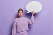 Unhappy sulking young European woman holds communication bubble for your advertisement wears casual knitted jumper and trousers looks discontent aside isolated over purple background thinks deeply