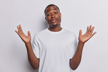 Indoor shot of hesitant dark skinned handsome man shrugs shoulders keeps palms raised up being doubtful dressed in casual t shirt isolated over white background. I dont know and cannot give you answer