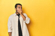 © Asier - Casual young Latino man against a vibrant yellow studio background, thoughtful looking to a copy space covering mouth with hand.